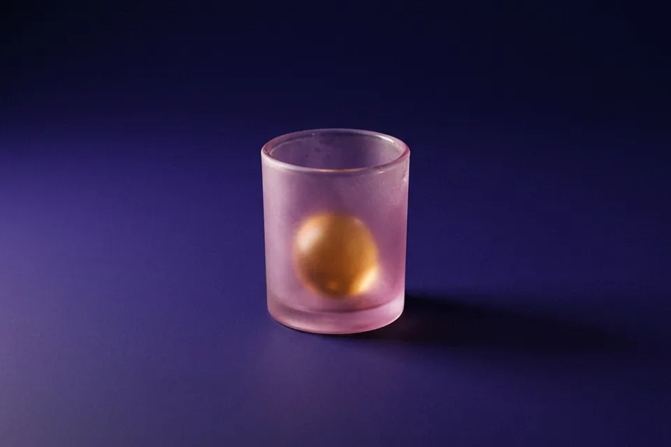 Esfera ámbar luminosa vista a través de un vaso de vidrio esmerilado rosado, sobre fondo violeta.