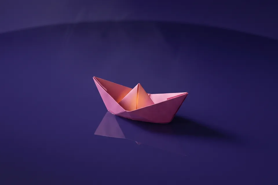 Barquito de papel rosa sobre una lámina de agua quieta que lo refleja, sobre fondo violeta.