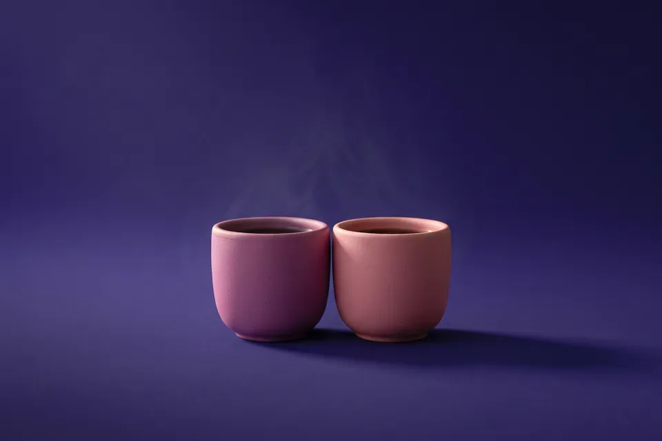 Dos tazas de cerámica, una magenta y otra rosa, juntas, con un hilo de vapor, sobre fondo violeta.